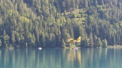Ein gelbes und rotes Löschflugzeug fliegt über dem türkisfarbenen Weissensee mit Booten und Bergen im Hintergrund.
