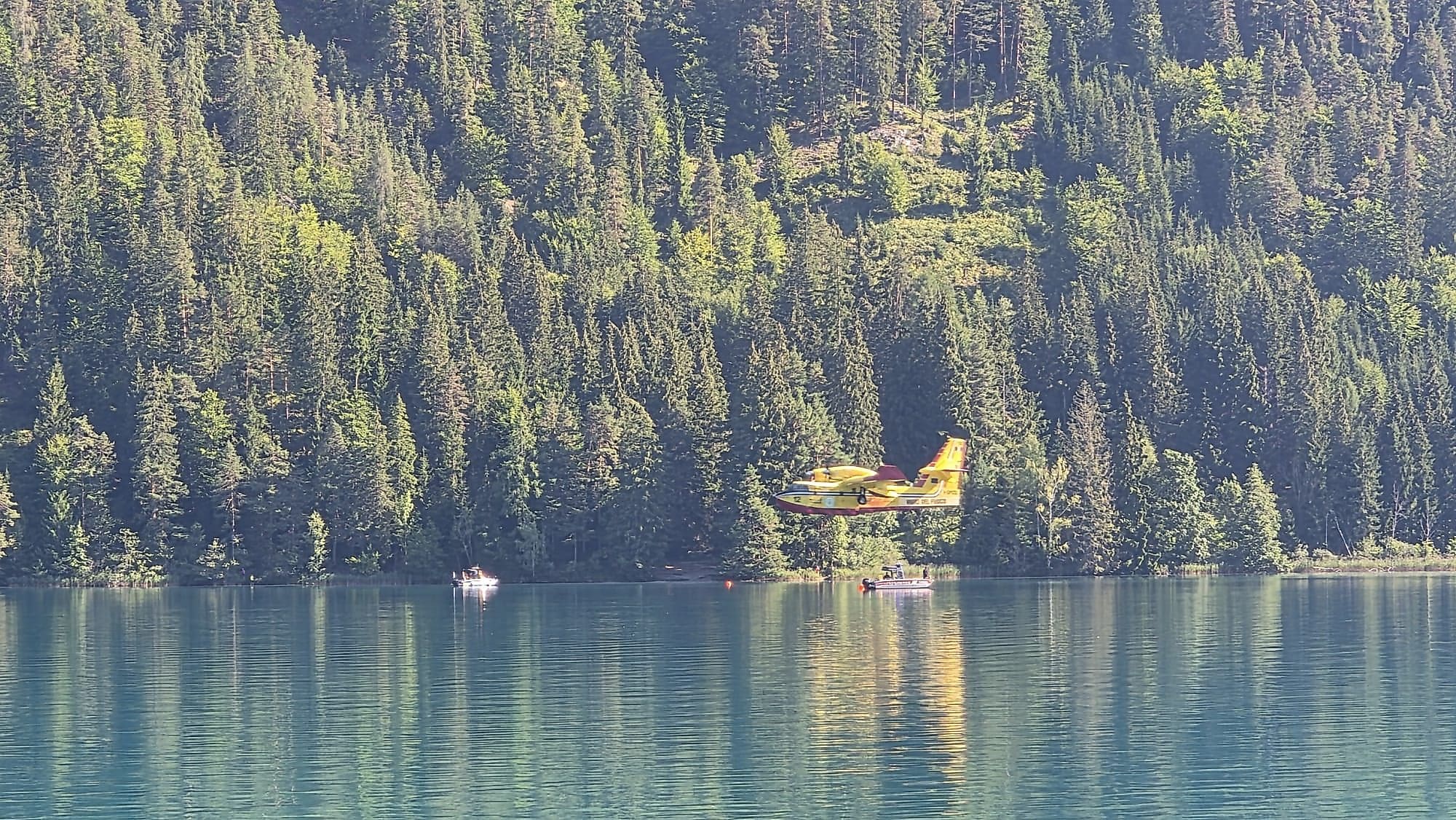 Ein gelbes und rotes Löschflugzeug fliegt über dem türkisfarbenen Weissensee mit Booten und Bergen im Hintergrund.