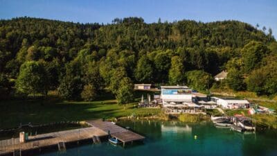 Die Luftaufnahme zeigt das Strandbad Maiernigg mit Holzstegen, Booten und einem Gebäude vor bewaldeten Hügeln.