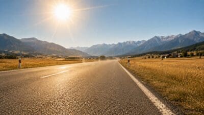 Eine Asphaltstraße führt durch ein sonniges Bergtal mit goldenen Feldern und Bergen im Hintergrund.