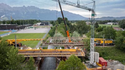 Bauarbeiter montieren Stahlträger einer Brücke über einem Fluss mit großen Kränen.