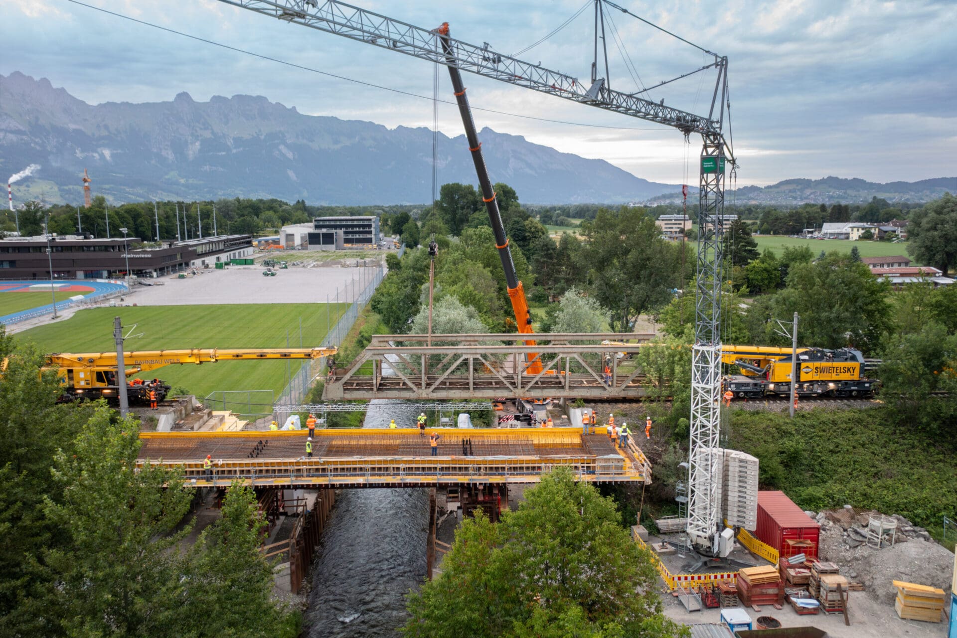Bauarbeiter montieren Stahlträger einer Brücke über einem Fluss mit großen Kränen.