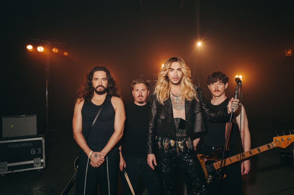 Vier Mitglieder der Band Tokio Hotel stehen auf einer dunklen Bühne mit Scheinwerfern.