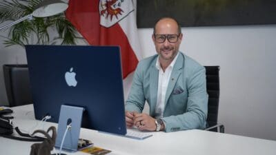 Ein Mann in einem hellblauen Anzug sitzt an einem Schreibtisch vor einem iMac und der Tiroler Flagge.