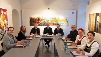 Gruppe von acht Personen in formeller Kleidung und Tracht an einem langen Tisch mit Dokumenten.