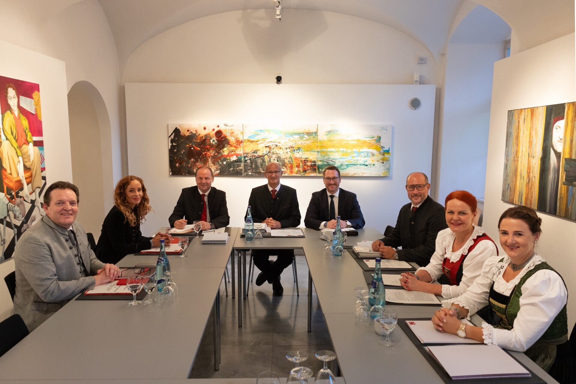 Gruppe von acht Personen in formeller Kleidung und Tracht an einem langen Tisch mit Dokumenten.