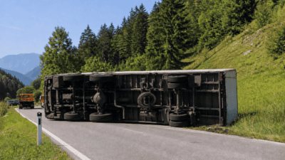 Umgestürzter grauer Lastwagen liegt quer über einer Bergstraße in bewaldeter Umgebung.