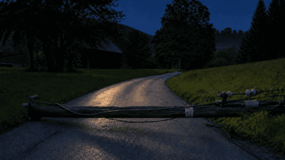 Umgestürzter Mast mit Leitungen quer über nasser Landstraße bei Dämmerung, umgeben von Bäumen und Wiesen