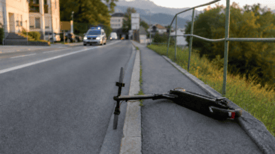 Schwarzer Elektroroller liegt umgestürzt am Bordstein einer Straße bei Dämmerung mit Polizeiauto im Hintergrund