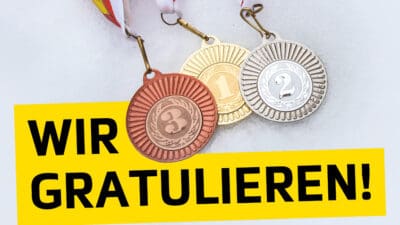Drei Medaillen aus Bronze Gold und Silber neben einem gelben Banner mit der Aufschrift Wir gratulieren