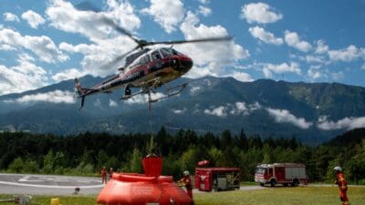 Polizeihubschrauber schwebt über großem orangenem Füllbecken mit Feuerwehrleuten in Tiroler Berglandschaft