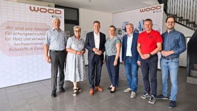 Sechs Erwachsene posieren für ein Foto vor großen Bannern des Wood K Plus in einem Eingangsbereich.