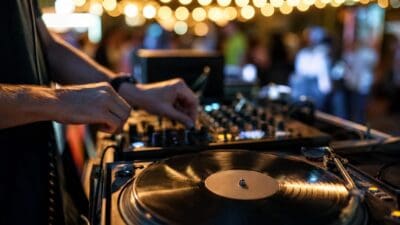 Nahaufnahme von DJ-Händen am Mischpult mit laufendem Plattenspieler und Lichterkette im Hintergrund