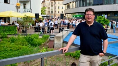Ein Mann im dunkelblauen Poloshirt lehnt mit der Hand am Geländer vor einer Menschenansammlung in der Stadt
