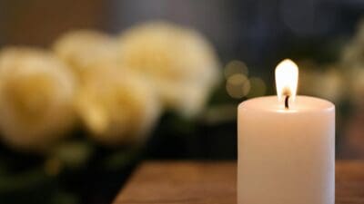 Eine brennende weiße Kerze steht auf einem Holztisch vor verschwommenen Blumen.