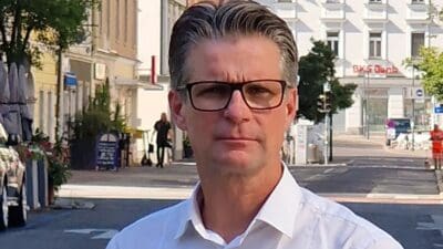 Ein Mann mittleren Alters mit Brille und weißem Hemd blickt ernst in die Kamera auf einer Straße.