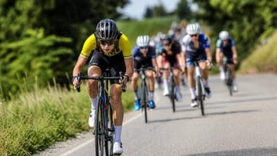 Radrennfahrer in gelbem Trikot mit Helm führt eine Gruppe von Radsportlern auf einer Straße an