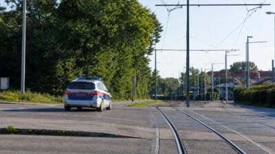 Ein silberner Polizeiwagen mit Blaulicht fährt neben Straßenbahngleisen unter grünen Bäumen.