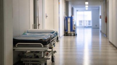 Ein leeres Krankenbett steht in einem langen Krankenhausflur mit Fliesenboden.