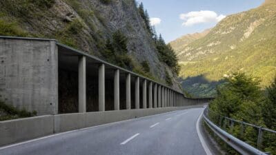 Kurvige Asphaltstraße verläuft neben einer langen Reihe von Betonpfeilern an einem steilen Bergabhang.