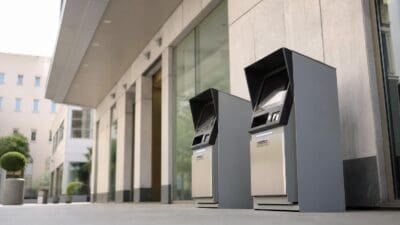 Zwei graue Bankomatkassen mit Bildschirmen vor einem Gebäude mit Glasfassade und Steinelementen