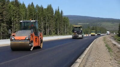 Eine orange Straßenwalze verdichtet frischen Asphalt auf einer neuen mehrspurigen Straße mit Wald im Hintergrund.