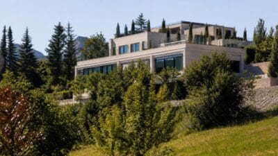 Mehrgeschossiges modernes Gebäude mit großen Fenstern und Terrassen inmitten von Bäumen vor Bergen