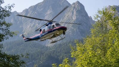 Polizeihubschrauber fliegt über einem dichten Wald vor einer Bergkette mit Felsgipfeln.