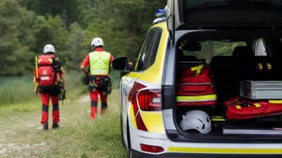 Zwei Bergretter in roter Kleidung gehen von einem gelben Rettungsfahrzeug mit geöffnetem Kofferraum weg