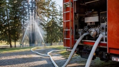 Rotes Feuerwehrfahrzeug mit offenen Türen und Schläuchen, im Hintergrund spritzen zwei Wasserstrahlen auf Bäume.