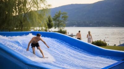 Ein junger Mann surft auf einer blauen mobilen Welle vor einem See mit Bergen im Hintergrund.