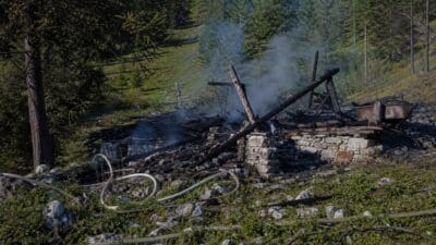 Rauchende Trümmer einer zerstörten Jagdhütte mit Feuerwehrschläuchen auf steinigem Boden im Wald