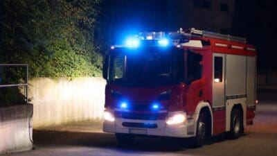 Frontansicht eines roten Feuerwehrfahrzeugs mit leuchtendem Blaulicht und Scheinwerfern bei Nacht.