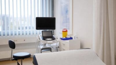 Ultraschallgerät steht in einem hellen Untersuchungsraum neben einem Monitor und einer Liege.