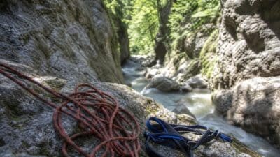 Aufgerolltes rotes Kletterseil und schwarzer Gurt auf moosbewachsenem Felsen in Schlucht mit Fluss