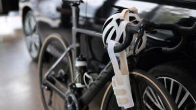 Ein Rennrad mit einem weißen Helm und einer beigen Handgelenksbandage am Lenker vor einem Auto.