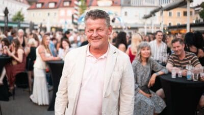Ein lächelnder Mann mittleren Alters in heller Jacke vor einer Menschenmenge bei einem Event.