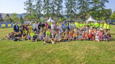 Große Gruppe von Kindern und Betreuern posiert auf einem sonnigen Sportplatz mit Zelten im Hintergrund