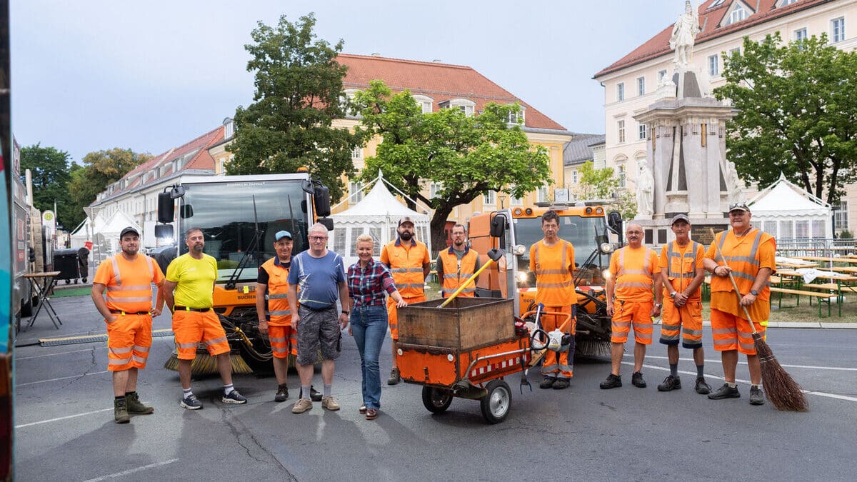 Gruppe von Reinigern in orangefarbenen Warnwesten mit Kehrmaschinen und einer Frau auf einem Stadtplatz
