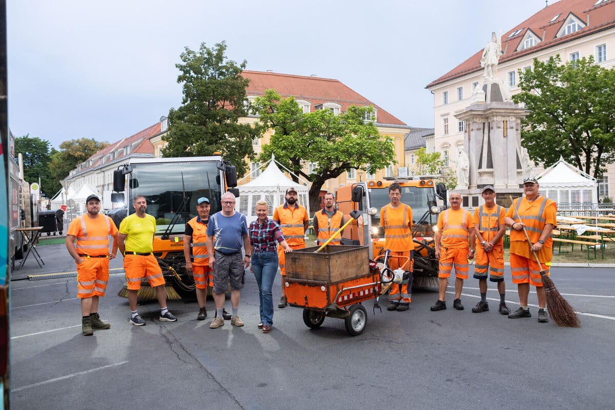 Gruppe von Reinigern in orangefarbenen Warnwesten mit Kehrmaschinen und einer Frau auf einem Stadtplatz