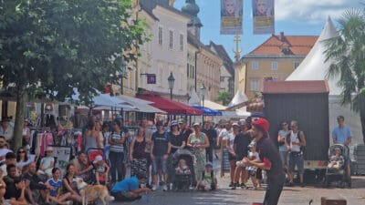 Straßenkünstler in roter Mütze tritt vor dicht gedrängtem Publikum auf einem Marktplatz in Klagenfurt auf