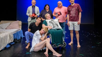 Schauspieler posieren auf einer Bühne mit blauem Hintergrund, Bett und Konfetti auf dem Boden.