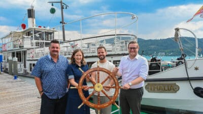 Vier Personen stehen auf einem Holzsteg und halten ein großes Steuerrad vor dem Schiff Thalia.