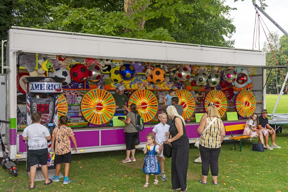 Besucher stehen vor einem bunten Spielstand mit Glücksrädern Plüschtieren und Fußbällen auf dem Festplatz
