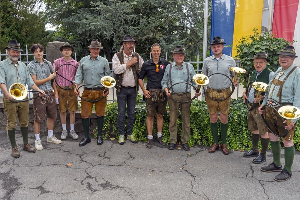 Männer in traditioneller Tracht halten Blasinstrumente vor grüner Hecke und Fahnen