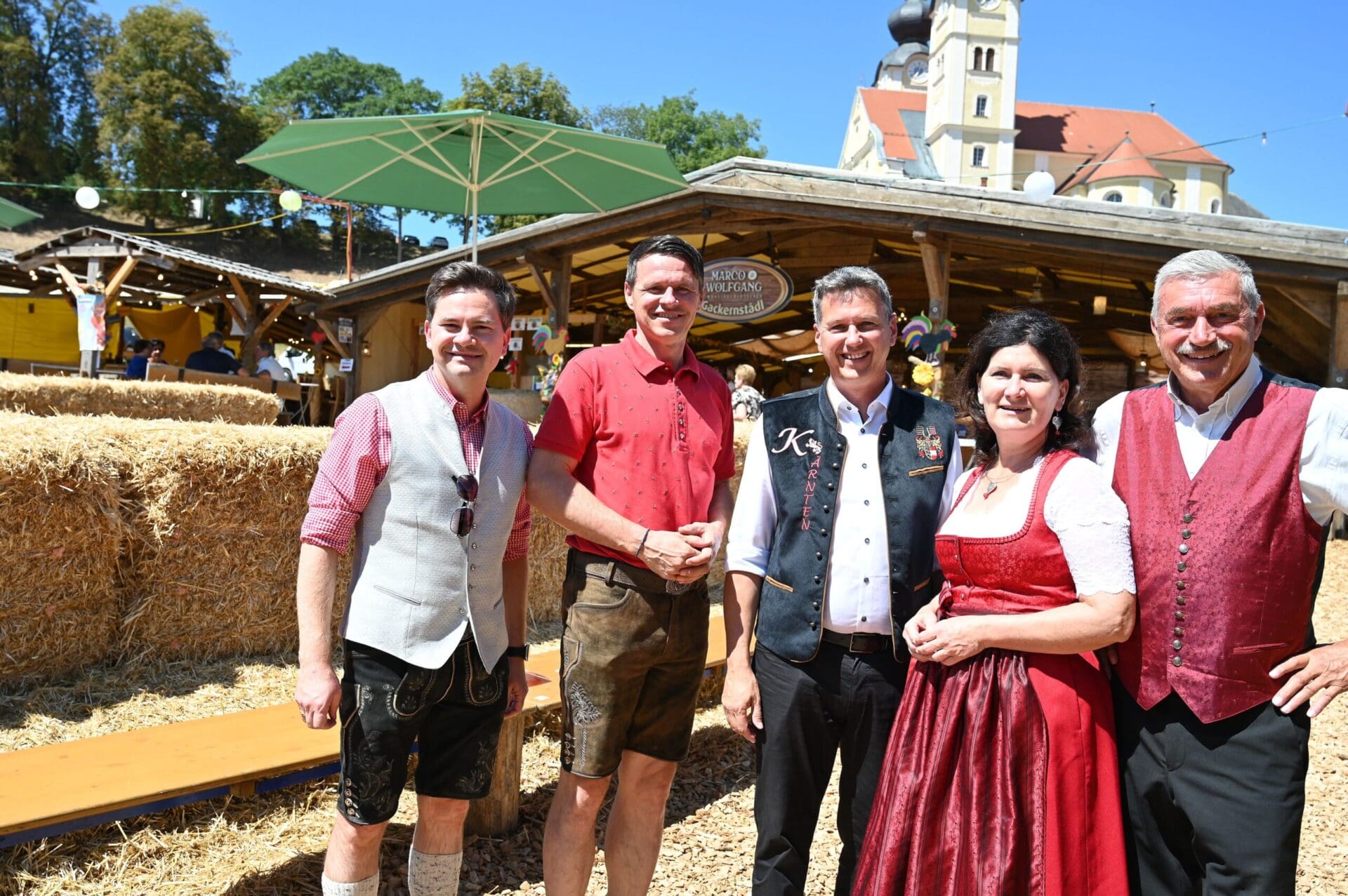 Fünf Personen in traditioneller Tracht stehen vor einem Festzelt mit Kirchturm im Hintergrund.