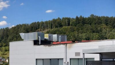 Weißes Betriebsgebäude mit Lüftungsanlagen auf dem Flachdach und sichtbaren Brandschäden vor bewaldetem Hügel