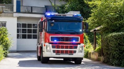 Rotes und weißes Feuerwehrfahrzeug mit eingeschalteten blauen Blaulichtern vor einem Gebäude.