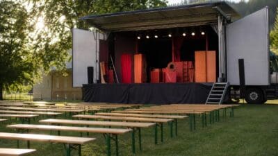 Offene Bühne eines Theaterwagens mit Bühnenaufbau und Reihen von Holzbänken auf einer Wiese.