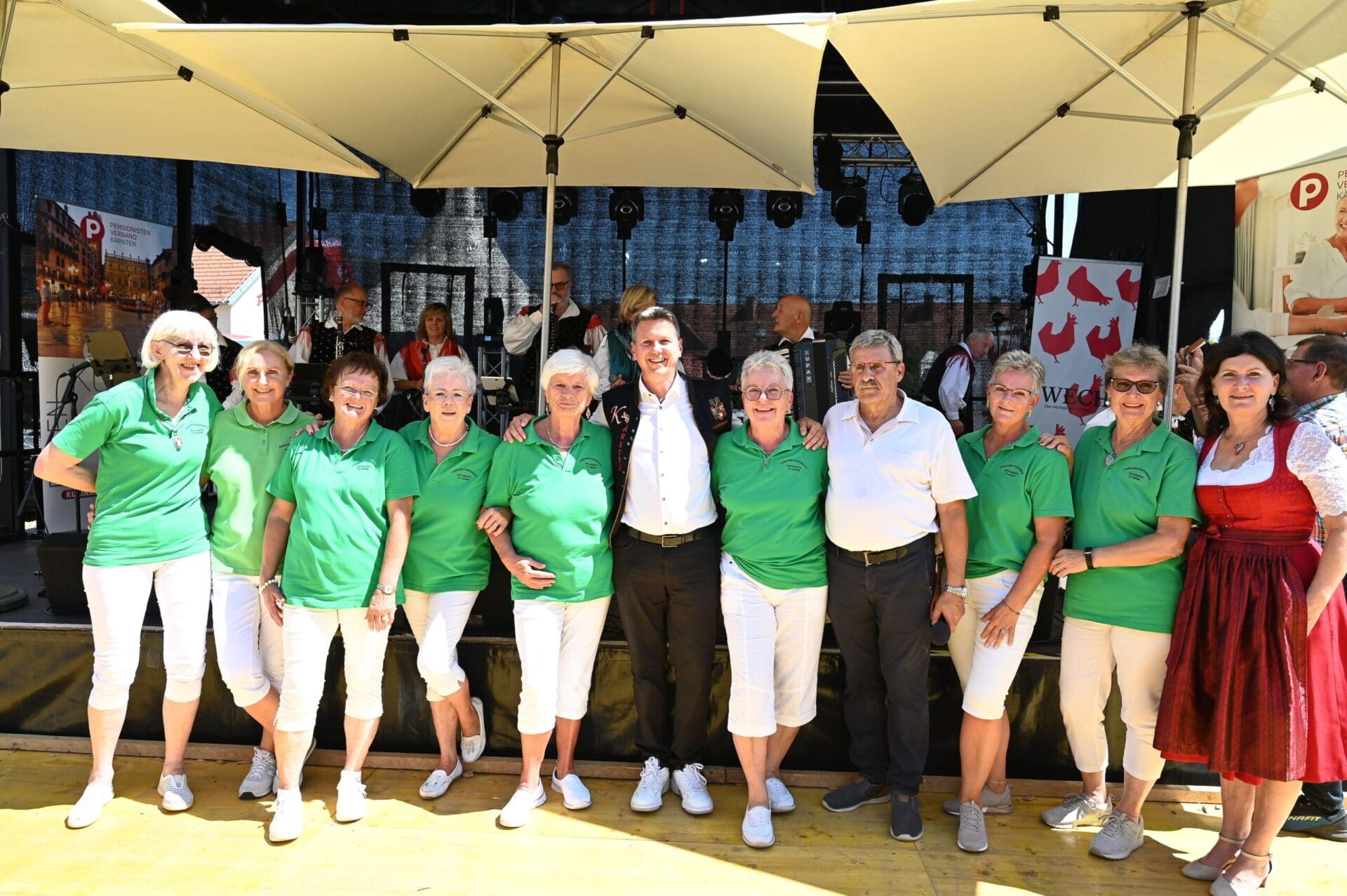 Gruppe älterer Frauen in grünen Shirts und ein Mann in weißem Hemd unter Sonnenschirmen auf einer Bühne